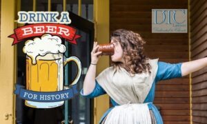 Woman in an 18th century blue dress with a white fichu over it drinks beer from a ceramic mug.